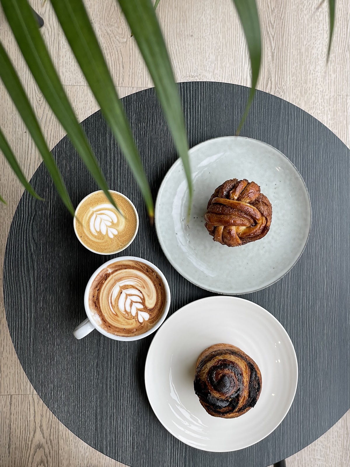 Seen from above on a black round table: a cardamom knot bun on a pale green plate, a chocolate swirl bun on a white plate, a cortado and a cappuccino, with palm leaves across the frame.