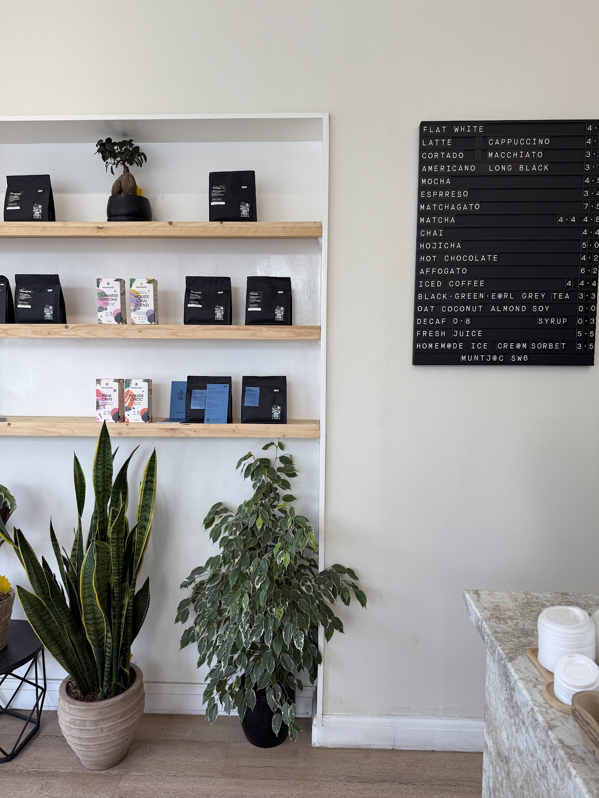 The black letterboard drinks menu on the wall of the shop, beside pale wooden shelves holding bags of coffee, Japanese matcha and house chai, with two large houseplants below.