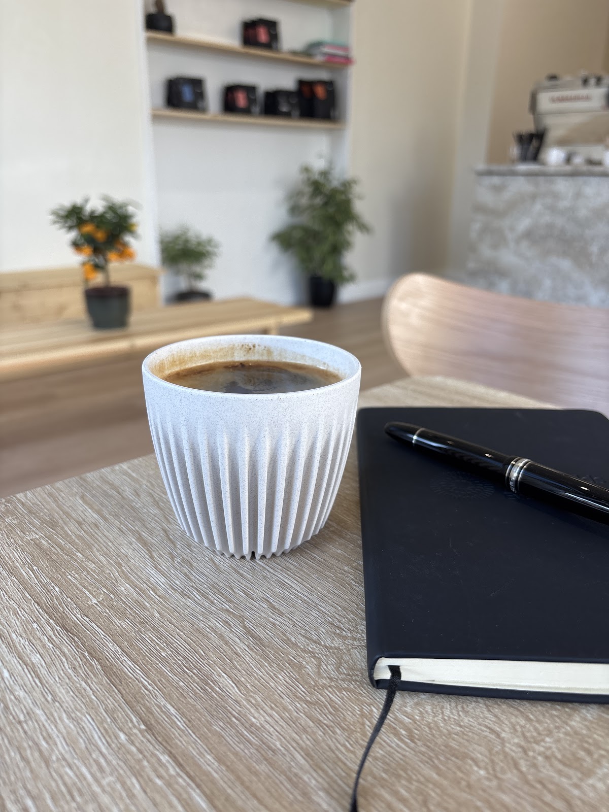 A black coffee in a ridged cream cup on a wooden table beside a notebook and pen, with the bright room, the retail shelf and the espresso machine behind.