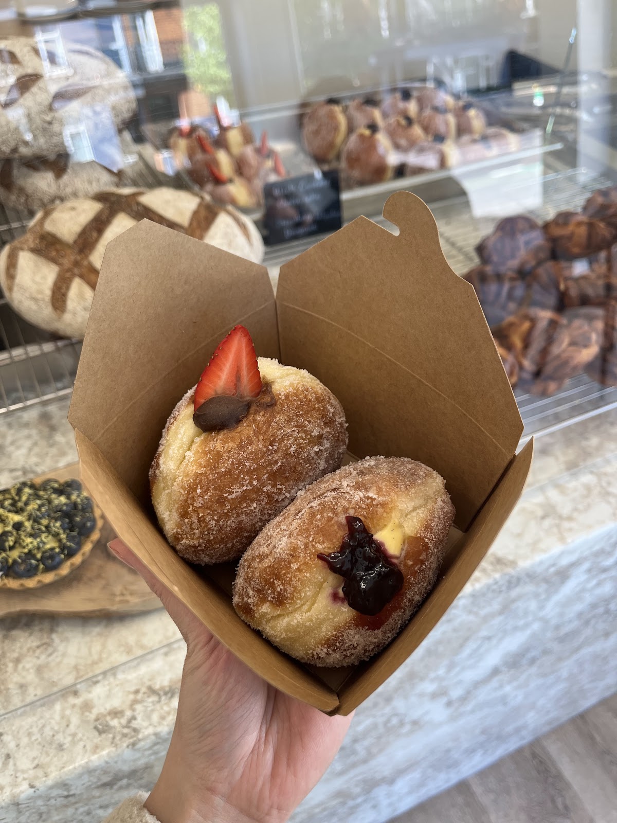 Two sugared doughnuts in a kraft takeaway box held up in front of the display case — one topped with a strawberry and chocolate, one filled with custard and dark jam.